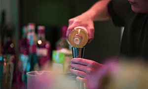 A bartender pours a drink into a jigger at a stylish bar with bottles in the background.
