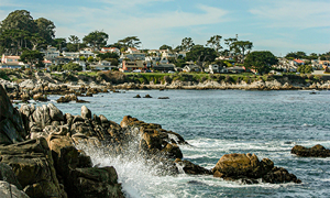 Waves crashing against rocky shore in Carmel-by-the-Sea with distant view of coastal town homes.