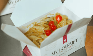 MV Gourmet takeout box with pasta and cherry tomatoes open on a kitchen counter.