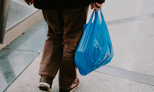 Rear view of a man carrying groceries in a blue plastic bag on an urban sidewalk.