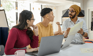 A group of adults engaged in a collaborative work discussion around laptops in a modern office setting.